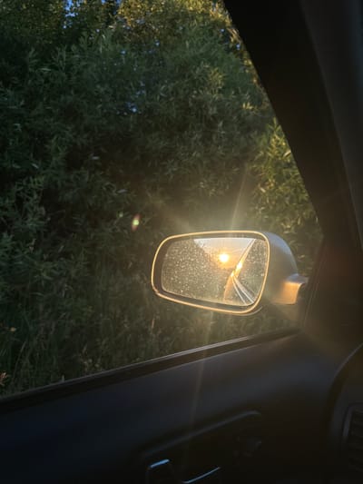 The image captures a view from inside a car, looking out the passenger-side window. A side-view mirror reflects a bright, setting sun through trees. The mirror is covered in small water droplets, suggesting recent rain or high humidity. Outside the window, dense green foliage with some dry grass is visible, illuminated by the warm light of the setting sun. The interior of the car, specifically the door panel, is dark and partially visible in the foreground, showing a door handle and window controls. There are no people visible in the image. The scene is likely from a rural or semi-rural area due to the abundant vegetation, and given the context of Tovstoluh, Ukraine, it depicts a typical roadside view at sunset in the region.
