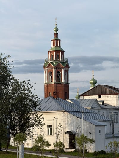 The image shows a brick bell tower with a green dome topped by a golden cross, characteristic of Russian Orthodox churches. The tower is partially obscured by a white building with a metal roof. In the background, another green dome with a cross is visible, suggesting the presence of a church complex. The sky is overcast with some patches of lighter clouds, indicating potentially diffused daylight. There are trees and green grass in the foreground and to the right of the buildings. The scene appears to be an exterior view of a historical or religious site. There is no visible text in the image. The orientation of the image suggests it was taken in portrait mode. The time of day is likely daytime, given the lighting.
