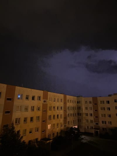 A nighttime aerial view shows a multi-story apartment building with numerous windows, some illuminated by interior lights, against a dark, cloudy sky. The building, an example of communist-era architecture common in Świdnica, Poland, is light-colored with orange accent panels on its facade. A few trees are visible at the base of the building. The sky appears turbulent with a hint of purple and white, suggesting stormy weather, possibly with lightning in the distance.