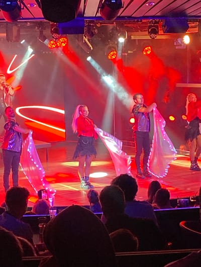 The image depicts a stage performance with several individuals on a brightly lit stage, predominantly in red and white lighting. In the foreground, the backs of an audience are visible, seated in what appears to be a performance venue.
On the stage, there are at least four performers. On the left, a male performer in a dark vest and black pants holds a microphone and appears to be singing. Next to him, a male performer in a dark vest and black pants is holding a flowing, sheer fabric that is illuminated with white lights. To the right of this performer, a female performer in a red jacket and a dark, distressed skirt stands holding a similar illuminated fabric. Further to the right, another female performer in a red outfit is visible, also holding a flowing fabric. The background of the stage features large, illuminated red shapes and fog or smoke effects.
The overall scene suggests a dynamic and visually striking performance. The audience is dimly lit, focusing attention on the illuminated stage. The presence of a microphone and varied costumes indicates a musical or theatrical performance. The location is described as the city of Hitis, Finland.