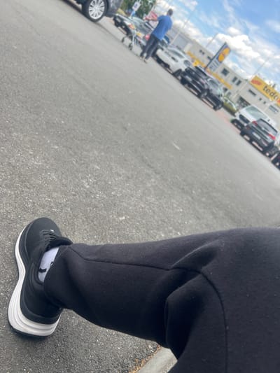 The image presents a low-angle perspective across an asphalt parking lot, leading towards commercial buildings under a partly cloudy sky. In the immediate foreground, positioned in the lower left, is the right leg and foot of a person. The person is wearing black sweatpants, white crew socks, and a black athletic shoe with a prominent white sole. The foot is resting on the coarse grey asphalt surface.
In the midground, several vehicles are parked. On the far left, the rear wheel and lower body of a silver SUV are visible. Further into the background, a person wearing a blue shirt and dark pants is seen pushing a metal shopping cart from left to right. Beyond them, a white sedan and multiple dark-colored cars are parked in rows.
The background features a commercial building complex. A tall signpost displays the yellow and blue "Lidl" logo, with "Lidl" and "PETERS" text underneath. To the right of the Lidl sign, another building exhibits a yellow and red "tedox" sign. More parked cars, including a white vehicle and a dark vehicle with its rear facing the viewer, are visible in front of these buildings. The sky above is blue with scattered white cumulus clouds.
Based on the visible "Lidl" and "tedox" establishments, the location is identified as a commercial area in Herne, Germany, specifically corresponding to the vicinity of Am Großmarkt 1, 44653 Herne.