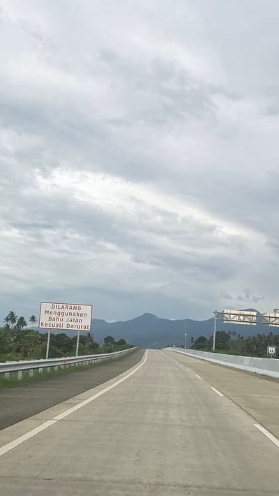 The image depicts a concrete highway in Baksukon, Indonesia. The sky is overcast with a cloud cover. In the distance, a mountain range is visible. A sign is located beside the highway with Indonesian text. The concrete highway has a white edge line. The location appears to be outdoors in daylight.