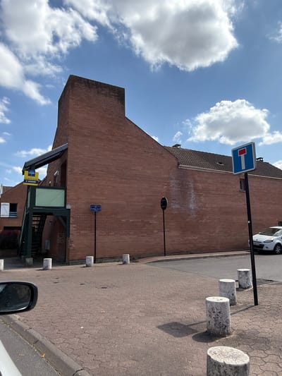 This is a vertical, eye-level, outdoor photograph taken on a sunny day with some clouds in a blue sky. The scene depicts a street in Évry-Courcouronnes, France, with a brick building on the left and a paved sidewalk with cylindrical concrete bollards on the right.
The building appears to be a two-story structure with a brown brick facade and a darker tiled roof. A sign with a blue background and a white horizontal bar is mounted on a pole in front of the building, indicating a dead-end street. Below that, another blue sign with white text is visible on the building. A green awning covers an entrance to the building, and a yellow real estate sign reading "À VENDRE" (For Sale) is attached to it.
On the sidewalk, several cylindrical concrete bollards are lined up, serving as a barrier. The ground is paved with interlocking bricks. A white car is parked on the street behind the bollards. The overall atmosphere is calm and ordinary, suggesting a typical day in a residential or commercial area. The sunlight casts shadows from the bollards and the awning onto the pavement.
Visible text includes:
- "À VENDRE" on a yellow sign.
- A blue street sign with white text.
- A smaller blue and white sign on the building.
- A license plate on the white car, reading "F 887 PX".