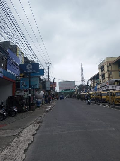 A straight road under an overcast sky is lined with various buildings and shops on either side. Utility poles and wires crisscross overhead. Motorbikes are parked on the left sidewalk, while yellow vans are parked on the right. A few people are visible along the sidewalk, and a scooter travels down the road. Several signs and advertisements are attached to the buildings, and a tall communication tower stands in the distance.