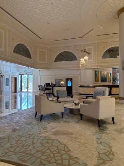 The image shows the interior of a hotel lobby in Ras Al-Khaimah, United Arab Emirates. The lobby features a high ceiling with intricate geometric patterns and decorative moldings. Natural light streams in through large glass doors at the far end, illuminating the polished marble floor which reflects the light and the surroundings. In the foreground, a seating area with light-colored armchairs and sofas is arranged around a circular coffee table with a glass bowl containing greenery. The furniture is placed on a large, patterned rug with abstract designs in shades of beige and light blue.
Further into the lobby, a reception desk is visible where a person is seated, seemingly attending to guests. Behind the reception desk, a curved counter with patterned panels is adorned with three abstract seascape paintings. Several decorative pendant lights hang from the ceiling above the seating area and the reception desk. To the left, a set of wooden doors and an exit sign are visible. The overall ambiance suggests a luxurious and modern hotel environment.