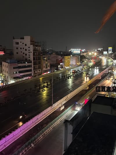 This image captures a nighttime view of a multi-level elevated highway in Shamshabad, India, likely during or shortly after rainfall. The wet asphalt reflects the bright headlights of numerous vehicles, including cars, trucks, and a bus prominently visible in the foreground. Buildings line the sides of the highway, some adorned with illuminated signage and decorative lights. One large billboard displays the word "BAWARCHI" in illuminated letters. The sky is dark and overcast. A distinctive orange flag is visible in the upper right corner of the frame. The scene depicts a busy urban environment with active traffic flow despite the nighttime and wet conditions.