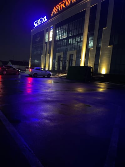 The image depicts a nighttime scene in Karmana, Uzbekistan, featuring a modern commercial building and a wet paved surface.

**Building:** A multi-story commercial structure dominates the background. Its facade features large glass windows, some reflecting interior light or external signage, and vertical architectural panels illuminated by upward-facing yellow floodlights. A prominent orange neon sign reading "NARVAN" is visible at the top, along with a blue neon sign reading "SaG XL". Cyrillic text, possibly "Цены от производителя" (Prices from the manufacturer), is visible on some windows. Two prominent entranceways with double doors are present at ground level, flanked by the illuminated panels. Scaffolding or temporary metal structures are visible near the left entrance.

**Ground:** The foreground and mid-ground consist of a wet, dark paved surface, likely a parking lot or street. This surface strongly reflects the artificial lights, creating prominent streaks of purple, blue, red, and yellow. A white painted line is visible on the left side of the paved area.

**Vehicles:** Two sedans are present. A white four-door sedan, equipped with a black spoiler on its trunk, is positioned in the mid-ground, angled slightly toward the left. Its taillights are off. Further back and to the left, a dark-colored (appears maroon or red) four-door sedan is partially visible, facing right, with its red taillights illuminated.

**Objects:** A dark green, rectangular object, possibly a dumpster or temporary barrier, is located near the right building entrance.

**People/Actions:** No individuals or overt actions are depicted in the scene.

**Environment:** The sky is dark and featureless, indicative of nighttime. Distant structures or trees are faintly visible on the horizon to the left. The overall environment is urban and commercially oriented, characterized by artificial illumination.