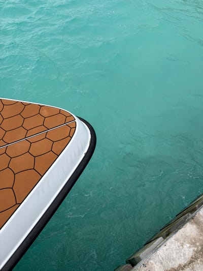 A high-angle shot shows part of a boat's bow adjacent to a concrete dock in a body of turquoise water. The boat features a brown honeycomb-patterned deck with black lines, bordered by a white rail and a black rub rail. The water is clear with subtle ripples. The concrete dock is weathered. The scene is devoid of people.
