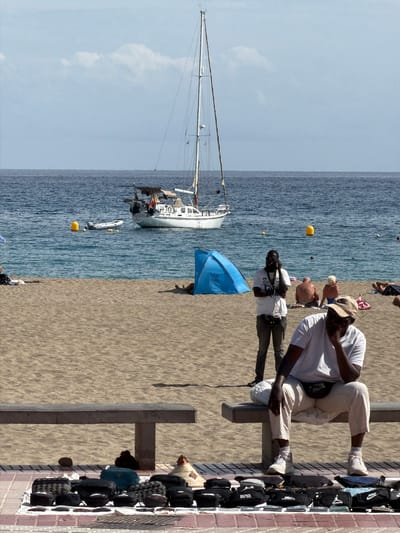 This image captures a vibrant beach scene in Los Cristianos, Spain, on a bright, sunny day. The clear blue sky is visible above a tranquil, blue-green ocean with small waves. A stretch of light brown sand occupies the foreground and mid-ground.
Several people are present, engaging in various activities. In the mid-ground, a man wearing a white t-shirt, light pants, and a baseball cap is seated on a low, concrete barrier or bench, possibly observing the scene or using a device. To his left, another man in a white t-shirt, dark pants, and sunglasses stands with his arms crossed, facing towards the water. A blue beach umbrella is partially visible behind him on the sand. Further in the background, other individuals are scattered across the beach and in the water, enjoying the sun and sea. One person near the water wears a blue swimsuit.
In the ocean, a large white sailboat with a tall mast is anchored on the left side of the frame, with a small dinghy or inflatable boat nearby. Two yellow buoys float in the water closer to the shore.
On the far right, alongside a paved walkway and a low brick wall, a display of merchandise is set up. Numerous bags, predominantly dark-colored fanny packs and larger bags, are laid out on white mats. Visible brand names on these items include "NIKE," "PUMA," "LACOSTE," and "BOSS." A vendor, partially obscured, appears to be attending to the display.
The time of day is likely midday, indicated by the high sun and distinct shadows. The overall atmosphere suggests a lively and typical beach day with both leisure and commercial activities taking place.