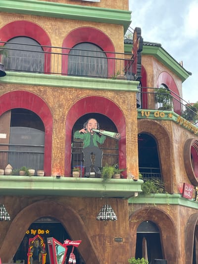 The image shows the exterior of a multi-story building, likely a restaurant or bar, with an earthy-toned facade featuring red arched windows and balconies. A prominent cutout figure of a person smoking what appears to be a large cigarette is positioned in one of the arched openings. Green trim runs along the edges of the building's levels.
On the upper right side of the building, text in yellow lettering is visible, which reads "TU DO" and "tu do trong". A red sign with white lettering that reads "QUÁN NHẬU" is also present.
Below, at the ground level, an arched entrance reveals neon signage. One sign spells out "TU DO" with stars on either side, and another sign is a Budweiser logo. The building is decorated with hanging light fixtures made of bottles. The overall setting appears to be a street in Nghĩa Trụ Commune, Vietnam, judging by the presence of Vietnamese text on the signage and the architectural style. The sky is overcast.