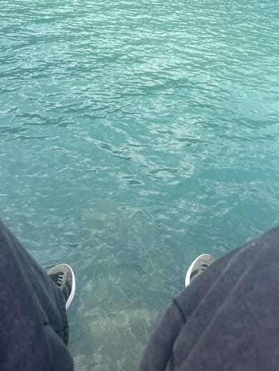 The image displays a first-person perspective, looking downward at a body of turquoise-colored water, likely Lake Brienz in Bönigen, Switzerland. The water's surface exhibits small, uniform ripples, and its clarity allows visibility of a light-colored lakebed directly beneath the observer's feet. In the foreground, the lower portions of two legs are visible, clad in dark grey or black casual trousers. Both feet are wearing grey and black athletic shoes with white soles; the left shoe shows black laces and a distinct white side stripe. The subject is positioned with their feet suspended directly over the water, indicating proximity to the lake's edge.