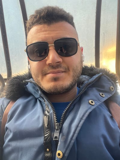 A male individual is depicted in a close-up selfie taken in Zarautz, Spain. He has short, dark, wavy hair and a trimmed beard, covering his chin and jawline. He wears aviator-style sunglasses with dark lenses and gold-colored frames, a white earbud in his right ear, and a blue hooded winter jacket with a dark faux fur trim around the collar. The jacket is partially unzipped, revealing a brighter blue long-sleeved shirt underneath and a prominent black zipper with a black, textured placket strip and brass-colored snap buttons. A brown strap, likely from a backpack, is visible over his right shoulder. Reflections of a smartphone, positioned vertically, are visible in the lenses of his sunglasses, indicating he is self-photographing. The background is blurred, showing an interior or sheltered space with a translucent panel on the right that permits a warm, orange-hued light to pass through, suggesting sunrise or sunset. A dark vertical structural beam is present on the left side of the frame.