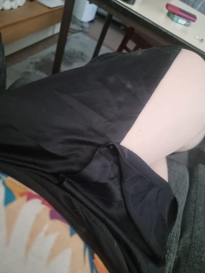 A close-up, high-angle shot shows a person's legs, draped in black fabric, likely a skirt or dress, resting on a gray sofa. The fabric is creased and appears to be made of a smooth, possibly satin material. A section of the person's bare leg is visible, extending from beneath the fabric. In the background, a wooden table with a white surface is visible, on which rest a pink object and a small green and white package, possibly cigarettes. The scene is indoors, with a carpeted floor and a wooden furniture visible in the background. The location in Carlisle, United Kingdom, is not discernible from the provided image.