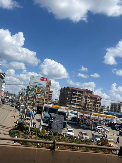 The image depicts a busy urban street scene in Kahawa Sukari ward, Kenya, viewed from an elevated vantage point. The sky is bright blue with scattered white cumulus clouds.
In the foreground, a concrete barrier is partially visible, behind which a dirt patch with sparse vegetation extends. A single individual, dressed in a white top and dark pants, walks on a dirt path in the lower-left.
The midground is dominated by a multi-lane road on the left, exhibiting moderate to heavy traffic consisting of numerous cars, vans, and public transport minibuses. A dense array of utility poles with overhead electrical wires lines the left side of the road. Further left, multi-story commercial or residential buildings are visible. One prominent building features a large billboard advertising "ZURI" and "KES 3M" with the text "BUILT FOR FAMILIES DESIGNED FOR GROWTH," and green signs below displaying phone numbers.
To the right of the road, a large Shell gas station operates under a yellow and red canopy, with multiple fuel pumps. Numerous vehicles, including cars, SUVs, and a blue truck, are present at the station, some appearing to be fueling. Several individuals are observed around the fuel pumps, functioning as attendants or customers. A large white sign with text is positioned near the gas station's entrance. Between the road and the gas station, an unpaved median strip with minimal vegetation is present.
In the background, multiple multi-story buildings, consistent with residential or commercial structures, are visible. One prominent building features a reddish-brown sloped roof and several visible balconies. The overall scene indicates active urban commerce and transit.