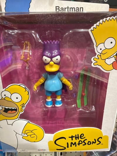 The media file shows a toy action figure of Bart Simpson, a character from the animated television series "The Simpsons," packaged for sale. The toy is inside a clear plastic blister pack, which is attached to a cardboard backing. Bart is depicted wearing his signature outfit: a blue shirt and blue shorts, with yellow arms and legs and white socks with blue stripes. He has purple spiky hair and large white eyes with black pupils. He is posed lying down within the packaging.
The cardboard backing features colorful illustrations of Bart Simpson and Homer Simpson. Bart is shown in a cartoonish, stylized representation on the left side of the packaging, with his arms outstretched. Homer Simpson is depicted on the right side of the packaging, with his mouth open in a surprised expression. The "The Simpsons" logo is prominently displayed on the right side of the packaging in yellow lettering.
Visible text includes "Bartman" vertically on the left side of the packaging, referring to one of Bart's superhero alter-egos. On the right side, the text "the Simpsons™" is in a large, stylized font. Smaller text that is partially visible includes "jakks" and "MATT GROENING". The background of the packaging is primarily a bright pink, with some purple elements in the toy's compartment. The overall scene is a retail product display, suggesting the item is for sale in a store. No specific time of day, weather, or explicit emotion beyond the characters' illustrated expressions is evident. The setting is implied to be a retail environment in Elizabeth City, United States of America, based on the provided location context.