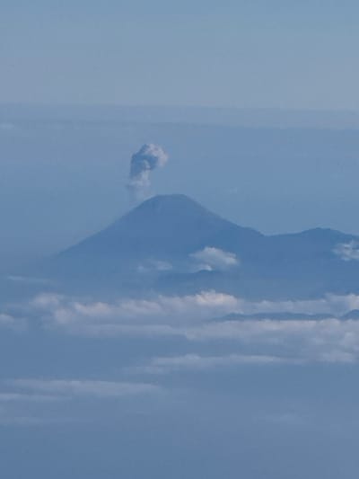 The image shows a volcanic eruption in a sky setting. A plume of ash and smoke billows from the peak of a volcano, which is partially obscured by clouds. The sky is a pale blue, suggesting it is daytime. The overall impression is one of raw natural power and the dynamic forces of the Earth. There is no visible text in the image. The scene depicts a moment of intense geological activity.