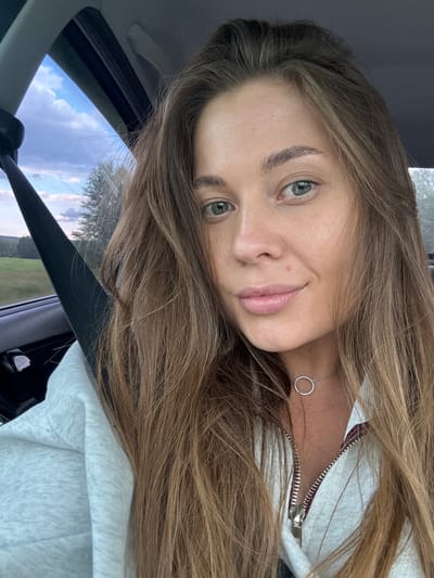 A close-up, selfie-style image shows a woman with long, light brown hair and green eyes. She is sitting in the passenger seat of a car, indicated by the visible seatbelt and car interior. The woman is wearing a light-colored, zippered hoodie and a delicate necklace with a circular pendant. The background outside the car window shows a landscape with trees and a cloudy sky, suggesting a rural or suburban setting, possibly on the outskirts of Бикбарда, Russia. The lighting is natural, suggesting it's daytime.