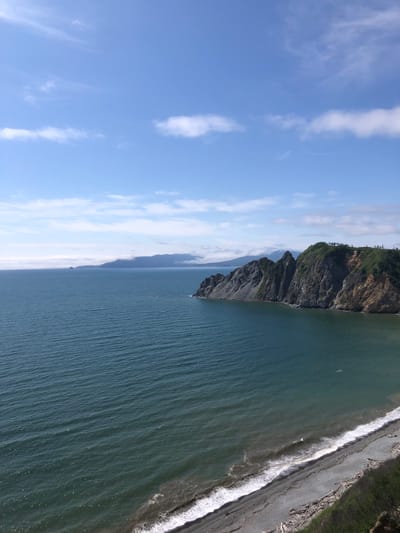 This is an eye-level, wide shot of a coastal landscape under a blue sky with scattered white clouds. The scene depicts a body of water, likely the ocean, meeting a rugged coastline.
The water occupies the majority of the left and center portions of the image. It appears dark blue-green with subtle ripples and waves indicating a moderate chop. The water's surface reflects the sky.
The coastline is characterized by steep, rocky cliffs that dominate the right side of the image. These cliffs are layered with grey rock and patches of green vegetation. One prominent section of the cliff face shows exposed soil and rock in earthy tones, suggesting erosion. The top of the cliffs is covered in dense green foliage and scattered trees.
A small, dark-colored beach with pebbles and scattered driftwood lines the base of the cliffs. White waves are breaking gently on the shore.
In the distance, a hazy mountain range or peninsula is visible across the water, shrouded in a light mist or fog. Two small, rocky islets are also present in the far distance. The overall impression is a remote and natural coastal environment. The location is identified as Ola, Russia.