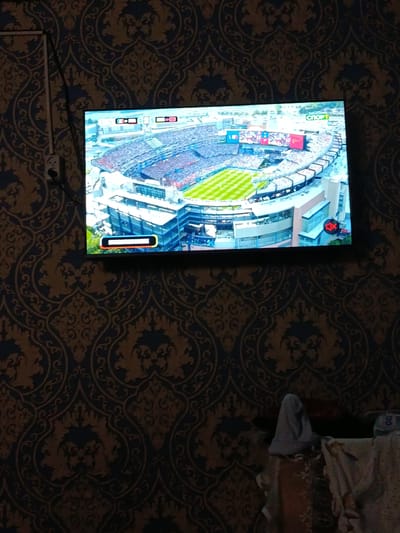A television screen displays a soccer match at a stadium. The game is broadcast on a channel with a logo that reads "СПОРТ" in Russian. The stadium is filled with spectators, and the field is green. A progress bar is visible at the bottom of the screen, indicating the current point in the broadcast. The television is mounted on a wall covered with dark blue and gold patterned wallpaper. To the left of the television, a white electrical outlet is visible. The overall environment suggests a domestic setting in an urban area, possibly an apartment or living room. The image does not provide any specific location information within the city of Кызыл-Октябрь, Kyrgyzstan.