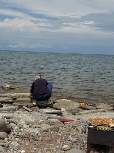 A person squats on a rocky shore of a body of water, facing away from the camera and towards the water. The person is wearing a dark t-shirt, blue jeans, and yellow shoes. In the foreground, a grill with cooked food is visible. The environment appears to be a natural setting with rocks of various sizes scattered along the shoreline and a calm body of water extending to the horizon under a cloudy sky. The location is the shore of Issyk-Kul Lake near Tong, Kyrgyzstan.