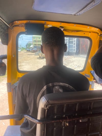 The image presents a forward-facing view from inside a yellow three-wheeled vehicle, likely a tricycle (keke napep), situated in Iyiowa Odekpa, Nigeria. The foreground is dominated by the back of an individual's head and upper torso, centrally positioned. This person has short, dark, closely-cropped hair and wears a dark-colored t-shirt, on the back of which a circular graphic or logo is partially visible. They are seated directly behind a black padded seat, which features a horizontal metal bar across its lower rear.
An electronic device, dark in color, is mounted above the vehicle's front windshield. To the right of the frame, a hand is partially visible, holding a smartphone with an illuminated screen.
Through the windshield, an unpaved, dusty road is visible, extending into the mid-ground. Several vehicles, including a light-colored sedan, are present on this road. On the left side of the road, a multi-story building with light-colored walls and green accents (possibly roofing or window frames) is discernible. To the immediate right, a darker-colored building with a ribbed or textured facade stands. The sky above is bright with some cloud cover, indicating daylight conditions, and sparse vegetation is visible on the horizon.