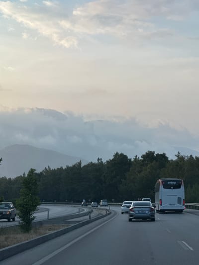 The image captures a highway scene in Döşemealtı, Antalya Province, Turkey, characterized by an overcast sky and mountainous terrain. The upper portion of the frame features a light blue sky transitioning into a dense layer of white and grey clouds that obscure the upper segments of distant mountains. Multiple layers of mountains, appearing hazy and grey-blue, are visible beneath the cloud cover. A dense, dark green forest occupies the midground, bordering the highway.
In the foreground, a multi-lane asphalt highway curves gently to the right. White lane markings are present. On the rightmost lane, a white passenger bus with "antvlp" displayed on its rear and the license plate "07 BPL 588" is visible. Directly behind the bus, a silver Honda Civic, bearing the license plate "07 CKN 617," is positioned in a central lane, followed by a white sedan. Further back, another light-colored vehicle is discernible. On a separate road or exit ramp to the left, an older model dark-colored car is present. Concrete barriers and guardrails line the road sections, with sparse vegetation and dry grass growing in the median. Yellow "K" shaped road signs, indicating a curve, are visible on the left side of the road. No individuals are clearly identifiable.