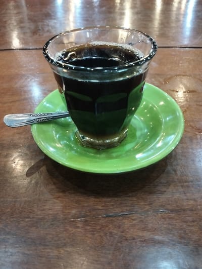 A clear, faceted glass containing a dark brown liquid with a thin layer of foam on its surface is centrally positioned on a glossy, circular, bright green saucer. A silver-toned spoon, featuring an ornate handle design, rests horizontally on the saucer to the left of the glass. This arrangement is set on a dark brown wooden table, exhibiting visible grain patterns, minor surface wear including scratches and a visible crack in the lower left portion of the frame, and reflecting multiple overhead light sources as elongated ovals and circles. The scene is located in Jeunieb, Indonesia. No individuals are visible.