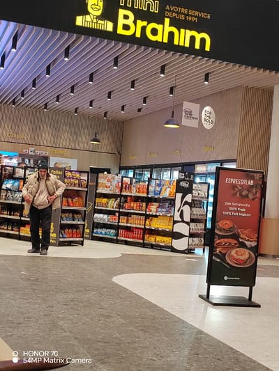 The image depicts the interior of a "Mini Brahim" store, "At your service since 1991." Shelves stocked with groceries and snacks line the aisles. A man stands near the "Tabac" section. Signage includes slogans like "Dare to sip different" and "For the bold." An "ESPRESSOLAB" sandwich advertisement highlights fresh, homemade sandwiches. The lighting is modern, with recessed lights illuminating the store. A "COOL COFFEE" display is also visible. A smartphone watermark "HONOR 70 54MP Matrix Camera" is present in the bottom left corner.