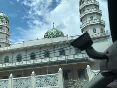 The image shows a view of a building identified as a mosque in Kruet Teumpeun, Indonesia, taken from inside a vehicle. The mosque features a large green-and-white patterned dome, two tall, multi-tiered minarets, and a facade with multiple windows and decorative elements. A low fence with geometric cutouts borders the front of the building. Utility lines are visible against the cloudy sky. A portion of the vehicle's interior, including a rearview mirror, is visible in the foreground, partially obstructing the view. No people are discernible in the image. The scene is captured during daylight hours.
