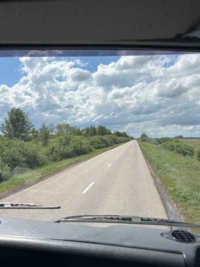 The image captures a view from inside a vehicle, looking out through the windshield onto an asphalt road stretching into the distance. The road is bordered by lush green vegetation, including trees and bushes. The sky above is filled with dramatic white cumulus clouds against a bright blue backdrop. The scene appears to be set in a rural or semi-rural area. The bottom portion of the image shows the vehicle's dashboard and windshield wipers. There is no specific information in the image that places it in Смирнов ауылдық округі, Kazakhstan.