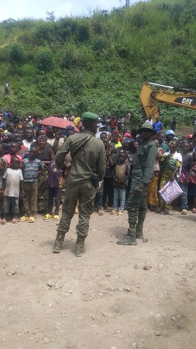 The image shows a gathering of people in Sake, Democratic Republic of Congo, at coordinates -1.57 latitude, 29.046 longitude. The scene is set in a dirt road with a steep, green hill in the background. An excavator, partially visible at the top of the image, is parked near the edge of the road. A group of people, mostly women and children, stand in a line facing the camera. Two men in military uniforms, one with a green cap and the other with a black hat, stand at the front of the group. They are looking straight at the camera. The people in the back are obscured by the group in front. The environment is dusty and dry with minimal vegetation. The people are dressed in various types of clothing, many wearing hats and head coverings. The scene suggests a community gathering or event.