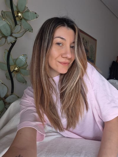 A female subject is depicted in a close-up, selfie-style indoor portrait, possibly within the Municipal Unit of Corinth, Greece. The adult female has long, straight brown hair with prominent blonde highlights, brown eyes, and is looking directly at the camera with a slight smile. She is wearing a light pink, short-sleeved t-shirt and appears to be reclining on a light-colored, textured blanket or bedding. A small, indistinct tattoo (possibly floral) is visible on the inner wrist of her left arm, which is extended towards the camera. To her left, a decorative lamp with a dark metal stem and two light-green, translucent, flower-shaped glass fixtures with brass centers is visible. In the upper right background, a framed piece of artwork, likely a tapestry with detailed figures and patterns, hangs on a light-colored wall. Partially visible to the far right are dark fabrics and a dark, indistinct object.