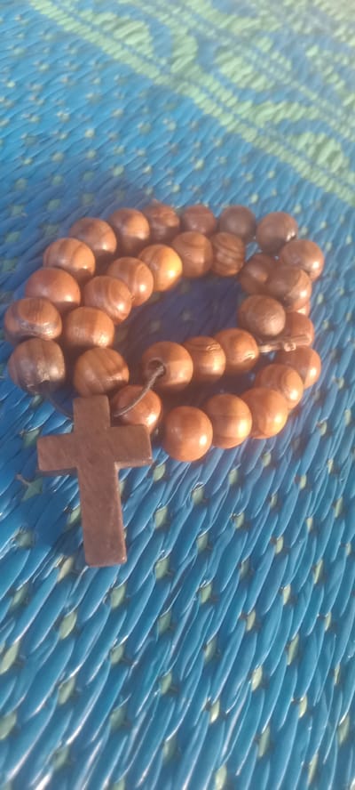 A wooden rosary with round beads and a cross lies on a textured blue and green mat. The scene is likely located in Bende, Nigeria. The rosary consists of smooth, rounded beads, strung together in a circle. A wooden cross is attached to the beads. The mat has a textured, woven pattern.