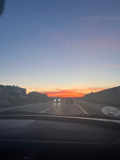 The image shows a view from inside a car driving on a road, likely near Palazuelos de Eresma, Spain, given the style of buildings visible. The sky shows a sunset gradient, transitioning from blue to orange and red hues. Other cars are visible on the road ahead, with their headlights illuminated. The car's interior elements are visible in the foreground, including the dashboard. Buildings line the side of the road.