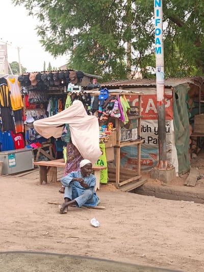 The image shows a street scene in Hotoro, Nigeria. An elderly man wearing a white skullcap and light blue clothing sits on the ground with his legs crossed and bare feet. He appears to be holding something small in his hand. Behind him is a small shop with clothing, hats, and other items on display. A woman stands near the shop, partially obscured by a draped white fabric. A sign reading "NIFOAM" is on a pole in the background. The ground is dirt, and there is a small piece of white trash nearby. There is a construction trench nearby. The shop sells clothes, hats, and possibly other small goods.