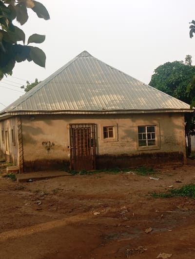 The image depicts a small, single-story structure with a conical metal roof, likely a dwelling or a small shop, situated in an outdoor environment. The walls of the building are made of a rough, light-colored material, appearing somewhat weathered and stained at the lower sections. The structure has a metal grated door in the center of the visible facade, flanked by windows. To the left of the door is a smaller, square window, and to the right, a larger rectangular window with white framing. Decorative vertical elements are present on the corners of the building. The ground in front of the structure is rough, unpaved earth, with some scattered debris, including leaves and small pieces of trash. Vegetation, including grass and what appear to be tree branches, is visible around the building and in the background. The sky is overcast or hazy. The overall impression is of a simple, possibly rural or semi-urban setting. The location is specified as Karsana 2, Nigeria.