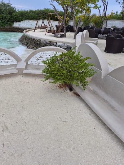The scene is an outdoor area in Bodufolhudhoo, Maldives. The ground is light-colored sand. A series of curved, white concrete structures define the space. A green shrub is positioned between two of these structures. In the background, there are dark-colored chairs and tables, suggesting an outdoor seating area. Further back, there's a wooden swing set. The structures lead to a pool of turquoise water, with a rock-lined edge and green vegetation on the far side. The sky is visible at the top of the frame.