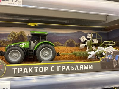 In Kuzmolovsky, Russia, a toy tractor with attached rakes is displayed on a shelf in what appears to be a retail environment. The toy tractor is green and grey, with large black tires and a visible engine compartment. It is connected to a green implement that resembles rakes, designed to collect hay or other agricultural materials. The toy is presented within its packaging, which features a scenic backdrop of a cultivated field and rolling hills. The packaging also has prominent text in Russian that translates to "Tractor with Rakes." The lighting suggests an indoor setting, likely a store aisle. There are no people or animals visible in the image.
