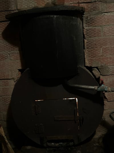 An old, black metal stove is positioned in front of a brick wall in Mel'nichnaya, Russia. The stove has a large, round base with a smaller, cylindrical component stacked on top. A pipe extends from the side of the stove and connects to a valve with a red handle. The stove's door is partially open, revealing a faint orange glow from within. A broom made of straw leans against the left side of the stove. The brick wall behind the stove is textured with a hexagonal pattern.