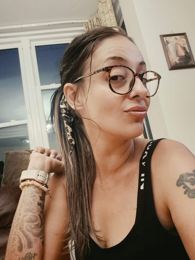 A woman is shown from the chest up, looking to her right. She is wearing glasses with brown tortoiseshell frames, a septum piercing, and a black tank top. Her hair is pulled back into a ponytail and has a leopard print accessory. She has a tattoo on her left arm and a watch and bracelets on her left wrist. A framed picture hangs on the wall behind her. The scene appears to be indoors.