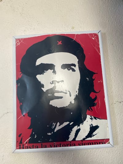 A framed poster depicting Ernesto "Che" Guevara in the iconic "Guerrillero Heroico" style is affixed to a textured, off-white wall in La Salle-les-Alpes, France. The graphic features a predominantly red background with Guevara's face, shoulder, and beret rendered in black and white tones. A stylized red insignia, resembling a 'K' or star, is visible on the beret. Along the bottom edge of the poster, the white text "Hasta la victoria siempre" is present. The poster, enclosed in a thin white frame, exhibits significant wear including numerous creases, surface abrasions, minor tears, and areas of discoloration, particularly across the red background and edges. The surrounding wall surface presents a granular, uneven texture with minor blemishes and fine linear cracks. No other individuals or objects are present within the captured scene.