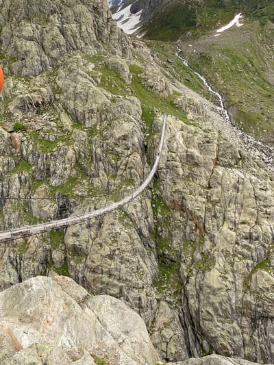 A high-angle, vertical shot depicts a rugged mountainous landscape in Innertkirchen, Switzerland, dominated by a long suspension footbridge spanning a deep valley. The bridge, constructed from wooden planks and metal cables, curves from the lower-left foreground towards the upper-right midground. Two small human figures are visible walking on the bridge: one closer to the foreground section and another further along its span. The surrounding mountain slopes are steep and rocky, characterized by grey-green rock formations interspersed with patches of vibrant green vegetation and sparse trees. In the upper-right background, a narrow stream or waterfall cascades down the rocky slope, with distinct patches of snow visible higher up the peaks. The scene captures a remote, high-alpine environment.