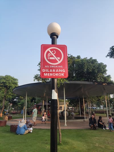 The image depicts a public park setting in Pagerwojo, Indonesia, under a clear blue sky. A prominent red sign with a white no-smoking symbol and the text "NO SMOKING DILARANG MEROKOK" is mounted on a lamppost in the foreground. The lamppost is black with white tape near its base and topped with a white globe light.
To the left of the lamppost, a woman in a blue hijab and a long-sleeved blue shirt sits on the grass with two children. One child is wearing a white hijab and a pink dress, and the other is wearing a white shirt and brown pants. They are facing towards the right of the frame.
In the middle ground, a covered pavilion with a dark roof supported by white pillars is visible. Several people are seated or standing within and around the pavilion. A man in a light green shirt and beige shorts walks away from the camera. Another person, possibly a child, wearing a white shirt and blue shorts runs towards the right.
To the right of the pavilion, a group of people are seated on the grass. Two women are squatting, one wearing a black long-sleeved shirt and patterned skirt, and the other wearing a black shirt and dark pants. A young girl in a pink t-shirt and blue jeans stands near them.
The park features well-maintained green grass, paved pathways, benches, and lush trees. In the background, a few buildings and a fence are visible. The overall scene suggests a relaxed afternoon gathering in a community space.