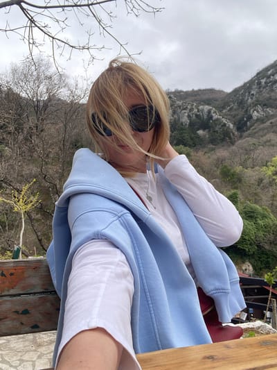 A photograph depicts a woman wearing sunglasses, a white shirt, and a light blue hooded sweatshirt. A small red purse sits next to her. She is seated on a wooden surface. Behind her is a landscape of trees and mountains, suggesting a natural setting. This scene is located outside of Krapina, Montenegro.