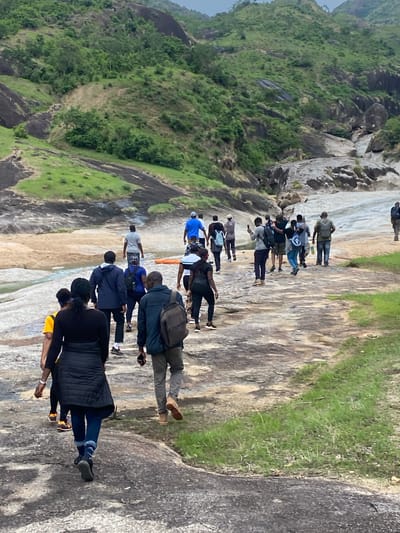 A group of approximately 18 people are standing or walking in a line on a large, rocky area. The rock is light colored and appears to be relatively smooth. To the left of the frame is a steep hillside covered in green vegetation. The sky is overcast. The people are wearing a variety of clothing. The scene appears to be in a natural, outdoor setting, most likely a rock outcropping or a mountainside location. It is in Ganawuri, Nigeria.