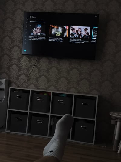 The image shows a television screen displaying a search results page for "Театр" (Theater) on Yandex. The results include several video thumbnails with titles such as "Карманный театр. Театр Ленком (1988)", "Закулисные войны. Театр", and "Цвет Малого театра в фильме-спектакле 'Дом Островского...'". The television is mounted on a wall with a patterned wallpaper. Below the television, there is a white shelving unit filled with dark gray storage boxes. The floor appears to be wooden. In the foreground, a person's leg and foot, wearing a white sock, are visible. The scene is dimly lit, suggesting it is likely evening or indoors with artificial lighting. The presence of a television and storage unit suggests a domestic setting. The location is identified as Rahachow, Belarus, although there are no specific visual cues in the image to confirm this location.