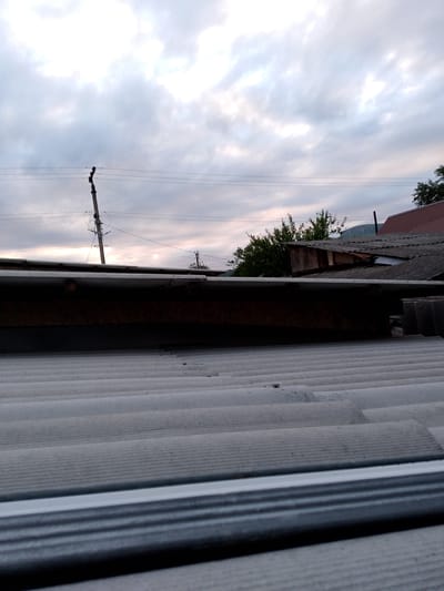 The image depicts a rural scene in Psebai, Russia, focusing on rooftops against a cloudy sky. The foreground is dominated by the corrugated texture of a light-colored roof, likely made of slate or metal. Above this, a darker, wooden structure is visible, possibly an attic or an extension. In the mid-ground, other rooftops are present, some appearing weathered and made of similar corrugated material, while others are tiled in red. Trees with green foliage are interspersed among the buildings. A prominent power line pole with insulators and wires stands tall, a common sight in rural areas. In the distance, a hilly landscape is faintly visible under the overcast sky.
The time of day appears to be late afternoon or early evening, suggested by the soft, diffused light and the muted colors in the sky. The weather is cloudy, with patches of brighter sky indicating the sun may be breaking through or setting. There are no people visible in the image. The overall mood is quiet and somewhat somber due to the overcast conditions. The setting is clearly a residential area, likely a village or small town, given the presence of houses and utility infrastructure. No text is visible in the image.