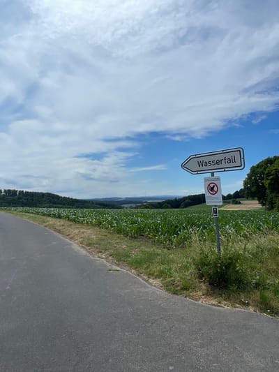 This is a landscape photograph taken in Christerode, Germany. The image features a road curving to the right, bordered by lush green grass and a field of crops. In the background, a dense forest stretches across rolling hills under a bright blue sky dotted with fluffy white clouds.
Dominating the left side of the frame is a white directional sign with a black border pointing to the left. The German word "Wasserfall" (waterfall) is written in black text on the sign. Next to it, on a separate pole, is a circular sign with a red border and a diagonal line across it, indicating prohibition. Inside the circle, a black silhouette of a dog is depicted, with text below it reading "Auf privaten Wiesen und Feldern..." which translates to "On private meadows and fields...". Another smaller sign with a black arrow pointing downwards is also visible on the same pole.
The scene suggests a pleasant daytime outing, possibly for hiking or cycling, given the road and directional signage. The weather appears to be clear and sunny, conducive to outdoor activities. The time of day is likely midday or early afternoon, based on the bright sunlight. The overall mood is one of tranquility and natural beauty, inviting exploration of the surrounding landscape.