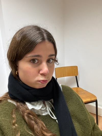 A female subject is captured in a close-up, frontal portrait indoors in Porto Salvo, Portugal. She possesses dark brown, wavy hair parted centrally, brown eyes gazing directly forward, and a pouting facial expression. Her right ear features a small gold hoop earring. She is dressed in a dark olive green top, layered over a light-colored, possibly checkered, collared shirt with white ties at the neckline, and a black scarf wrapped around her neck. In the background to the subject's right, a light brown wooden chair with a black metal frame and visible rivets on its backrest is positioned against an off-white wall corner. The lighting is even and consistent throughout the scene.