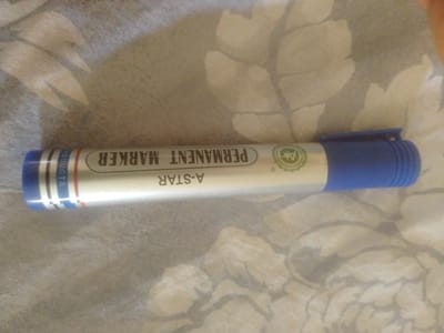 A blue and white permanent marker pen lies horizontally on a patterned fabric surface. The pen has the brand name "A-STAR" and "PERMANENT MARKER" printed in black on its white barrel. The fabric has a subtle floral or leaf-like pattern in shades of gray and beige. The image is a close-up shot, showing the entirety of the marker and a portion of the fabric. There are no people or specific landmarks visible that would indicate a location in Majemu, Nigeria.